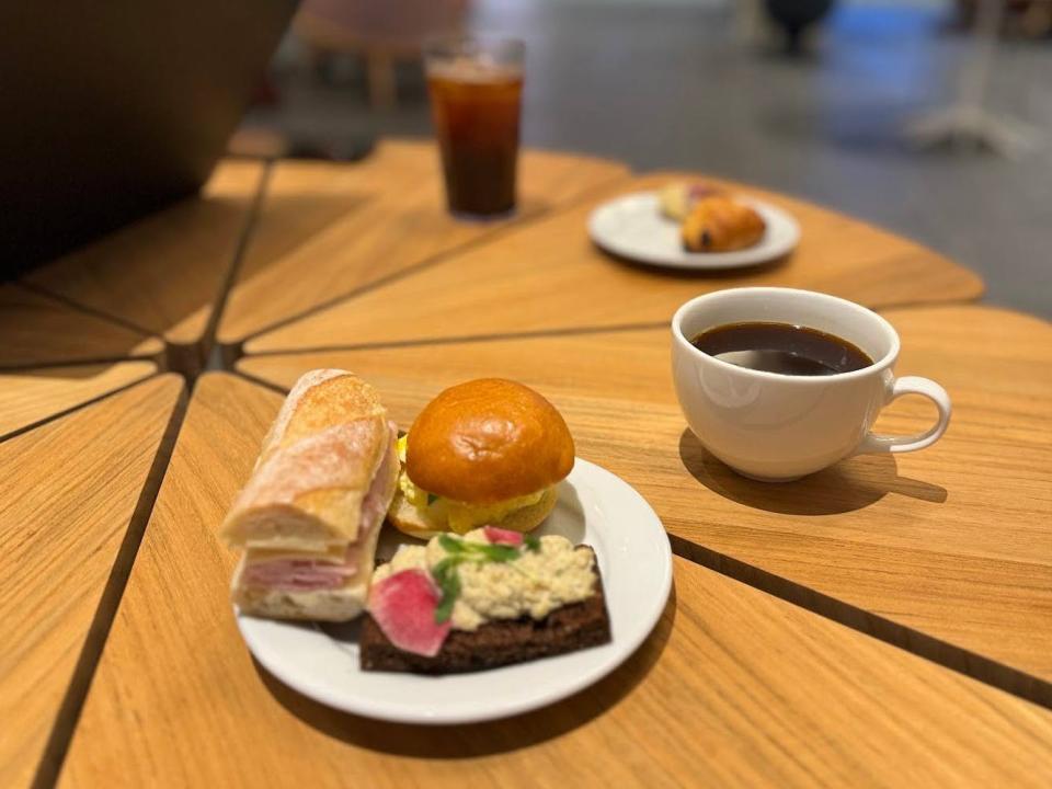 An Americano in a mug, and a mini egg salad sandwich, a mini ham and swiss sandwich, and a smoked lake trout tartine on a plate.
