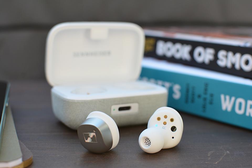 White/silver Sennheiser Momentum True Wireless 4 earbuds laying on a wooden table with the case open behind them. 