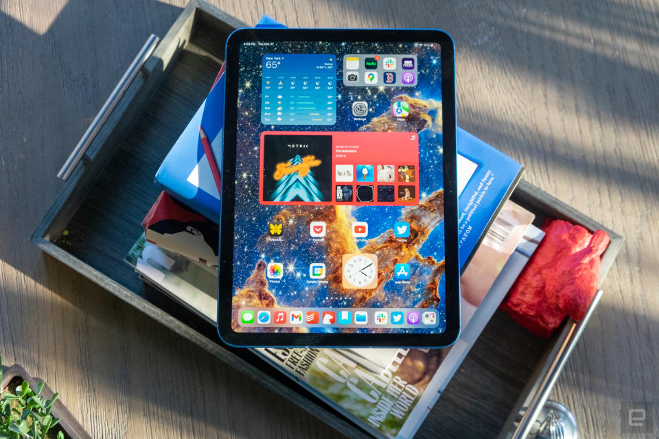 The 10th-generation iPad rests outside on top of a stack of books, on a wooden desktop, with its screen active, displaying a colorful home screen with various iPadOS widgets and apps.