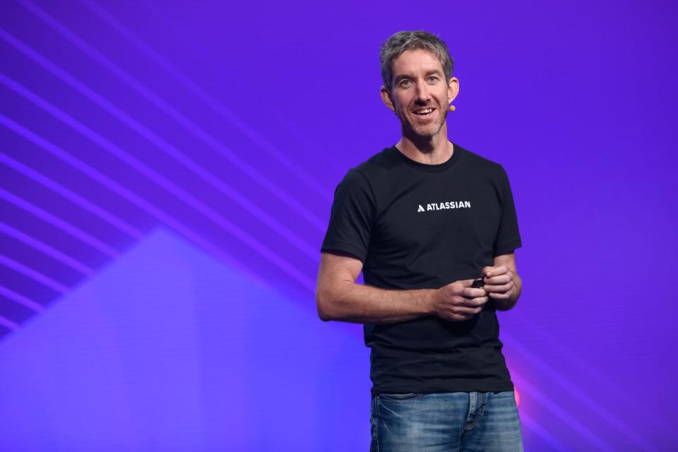 Scott Farquhar smiling on stage whilst wearing a microphone and Atlassian black t-shirt and jeans