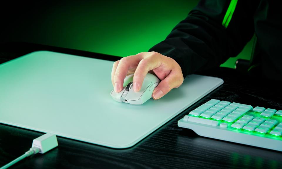 A white Razer Viper V3 Pro gaming mouse is held on top of a white mouse mat on a black desk, with a white keyboard glowing with green lighting situated off to its side.