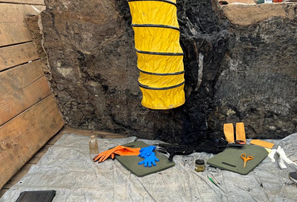 A large yellow tube hangs down in front of a large asphalt fossil deposit with tools on the ground