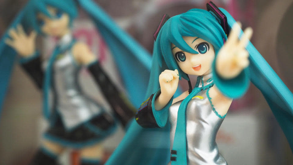 Closeup of a Hatsune Miku figure. She's waving a peace sign at the camera as a second figure in a different pose sits (blurred) behind.