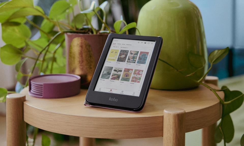 Product lifestyle photo of the Kobo Clara Colour ereader. The device sits propped up on a small, round, wooden table in front of vases with plants.