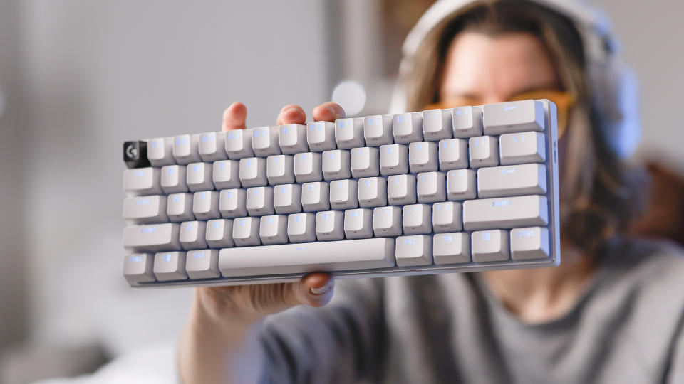 A woman in a gray sweater holds up a white Logitech G Pro X 60 gaming keyboard up to the camera with her left hand.