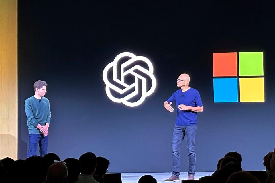 Two men on a stage — Sam Altman of OpenAI and Satya Nadella of Microsoft — with their corporate logos in the background, the OpenAI knott and MSFT's quad-colored squares