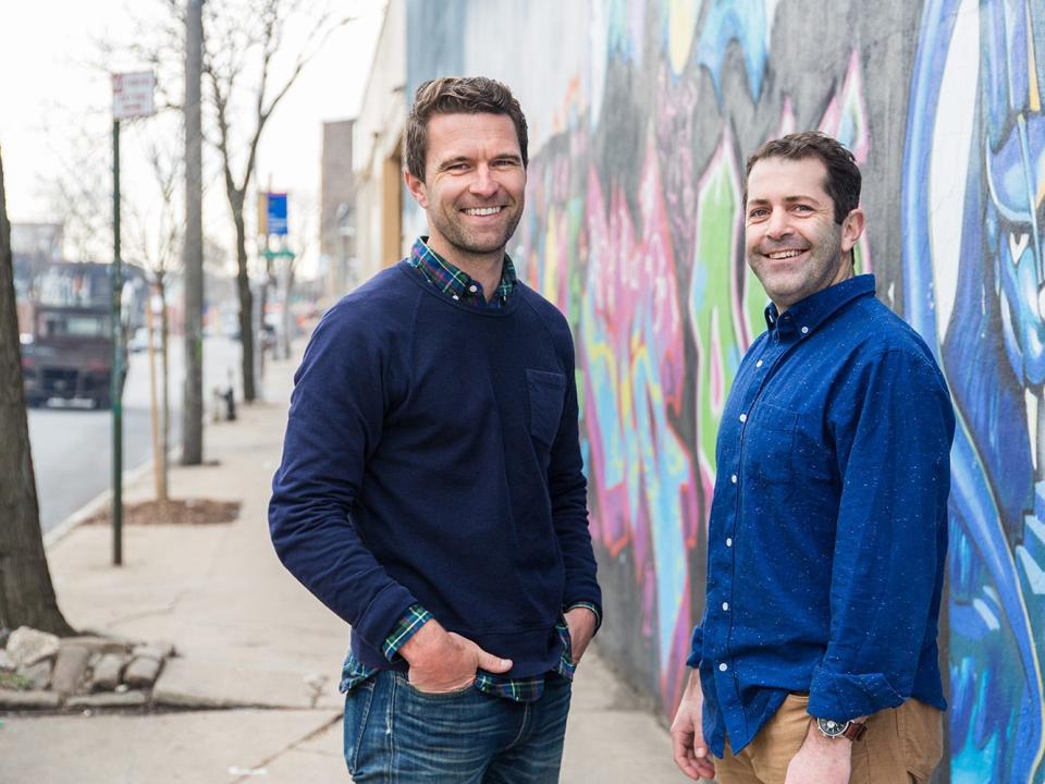Tim Brown and Joey Zwillinger posing for a photo against a graffitied wall on a sidewalk in an urban area/