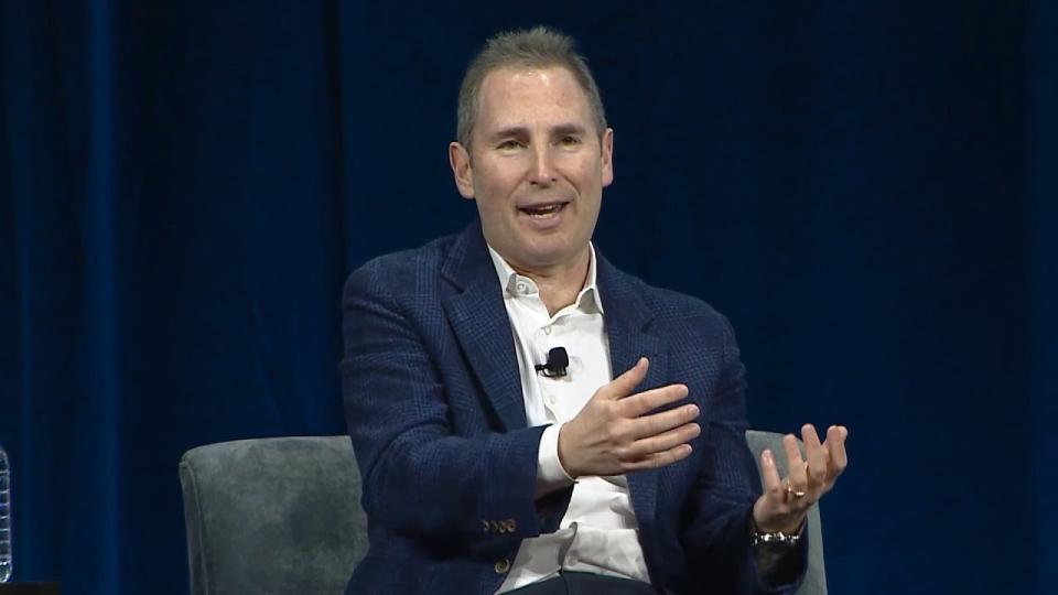 Amazon CEO Andy Jassy gestures toward the audience as part of an onstage talk. Black backdrop.